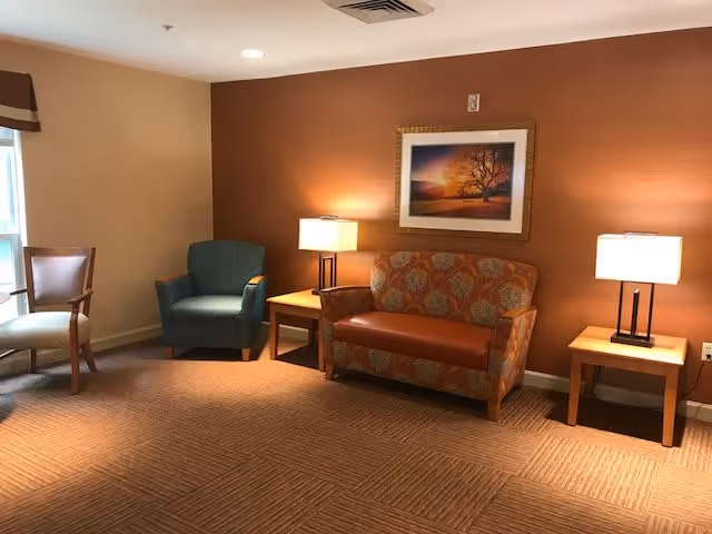 A cozy living room area with a patterned loveseat, a blue armchair, and a wooden chair near a window. Two wooden side tables with lamps flank the loveseat, and a framed picture of a tree at sunset hangs on the orange accent wall behind the loveseat. The carpet has a textured pattern, and the room is softly lit.