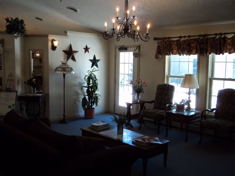 A cozy living room area with a dark sofa, two patterned armchairs, a wooden coffee table with magazines, and side tables with lamps and plants. A chandelier hangs from the ceiling, and decorative stars are mounted on the wall near a glass door letting in natural light.