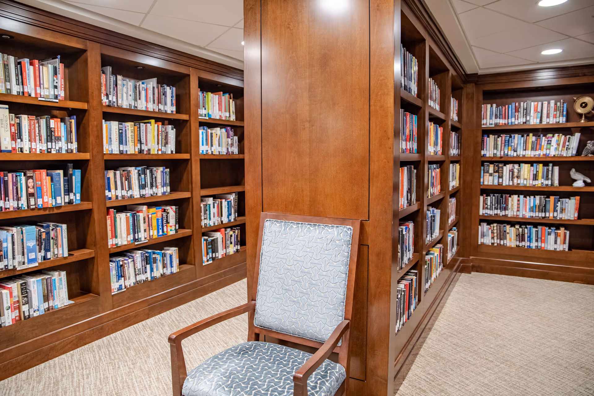 Interior view of a library room with wooden bookshelves filled with books lining the walls. A wooden chair with a patterned blue cushion is positioned in front of a wooden pillar. The room has a beige carpet and a white ceiling with recessed lighting.