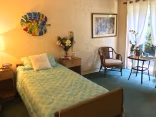 A cozy bedroom with a single bed covered in a light green quilt, a wooden nightstand with a lamp and flowers, a wooden chair, a small round table with a plant, and a window with white curtains letting in natural light. The walls are decorated with a colorful circular wall art and a framed picture.