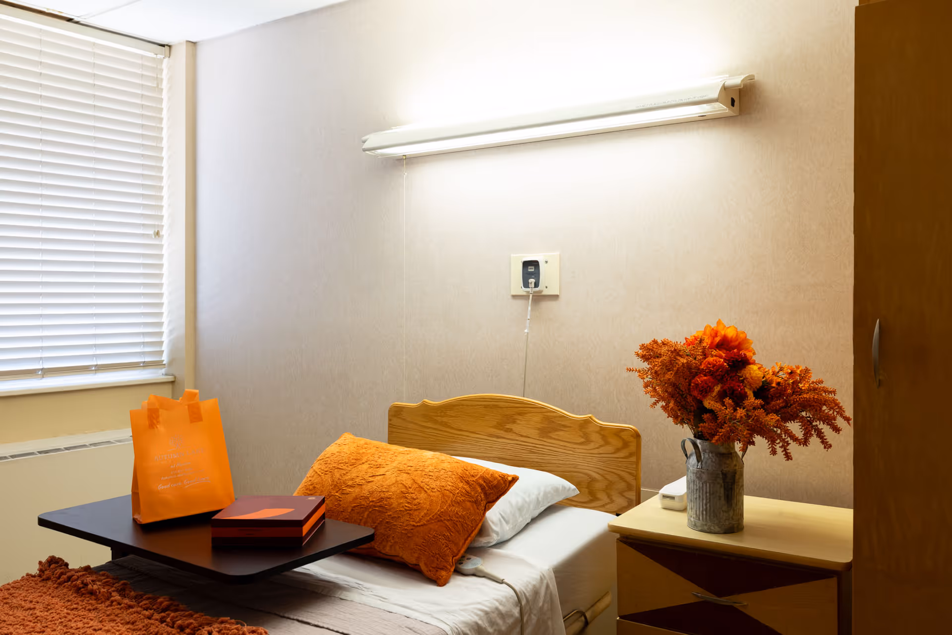 A single bed in a room with an orange pillow and blanket, a wooden headboard, a small bedside table with a vase of orange flowers, a window with closed blinds, and a black overbed table holding an orange gift bag and a box.
