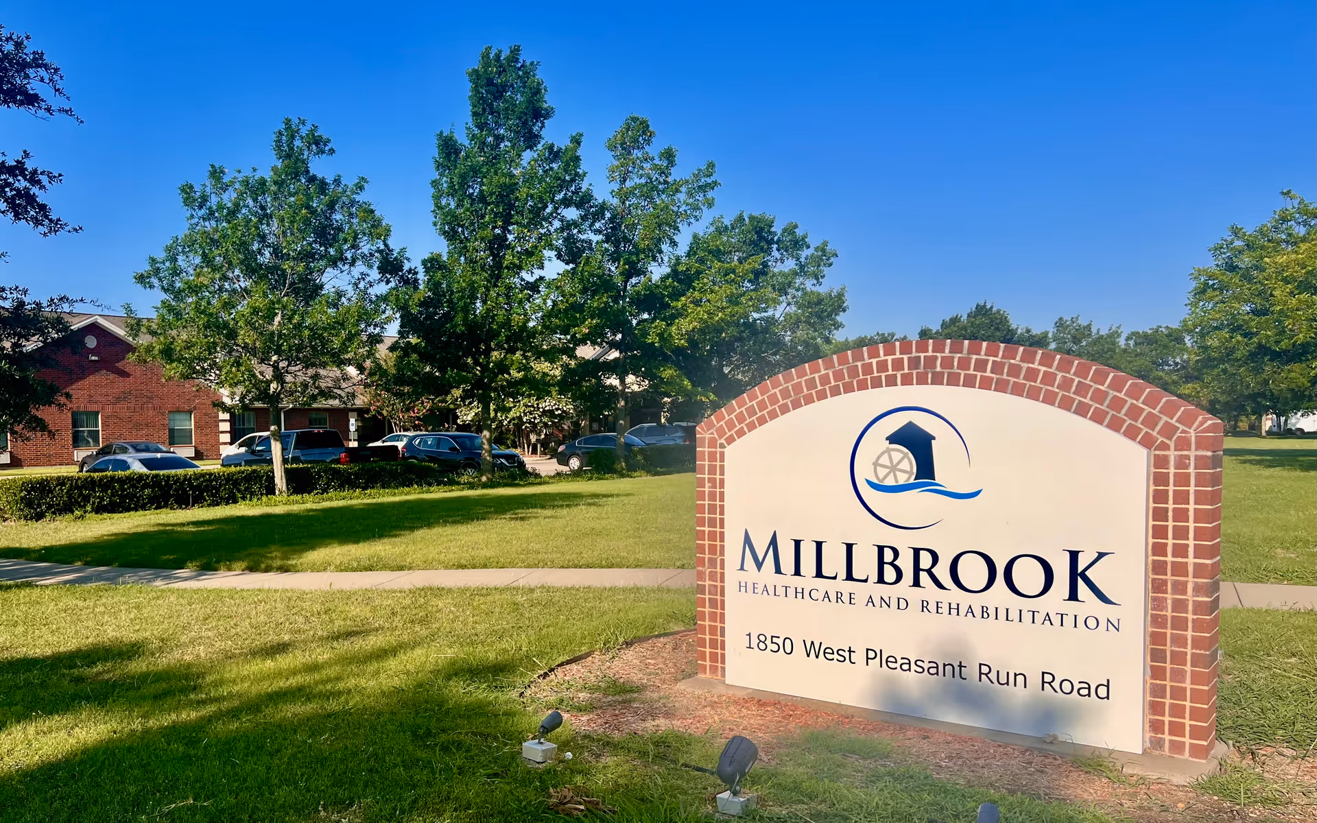 Outdoor view of Millbrook Healthcare and Rehabilitation Center sign on a grassy lawn with trees and a brick building in the background under a clear blue sky.