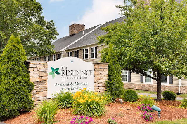 Exterior view of The Residence at Freeman Lake assisted and memory care living facility with a large sign in front surrounded by green bushes, colorful flowers, and trees. The building has beige siding, multiple windows, and a gray roof under a partly cloudy sky.