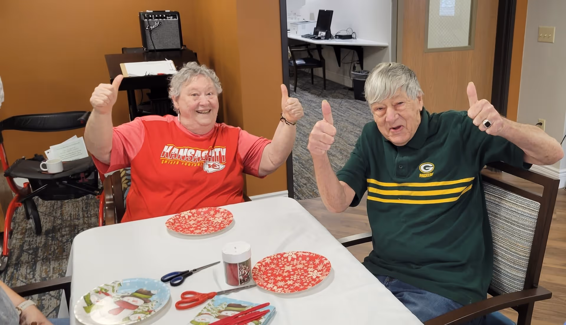 Two elderly people sitting at a table indoors, smiling and giving thumbs up. The person on the left is wearing a red Kansas City Chiefs shirt, and the person on the right is wearing a green Green Bay Packers shirt. The table has holiday-themed plates, scissors, and craft supplies on it. The background shows a brown wall, a black chair, and an open doorway to another room.