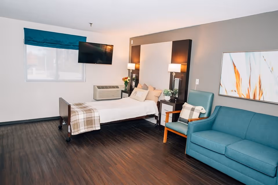 A clean and well-lit bedroom in a senior living facility featuring a single bed with white bedding and a plaid blanket, a teal armchair with a plaid pillow, a teal sofa, two bedside tables with lamps and plants, a wall-mounted TV, and a large window with a teal curtain.