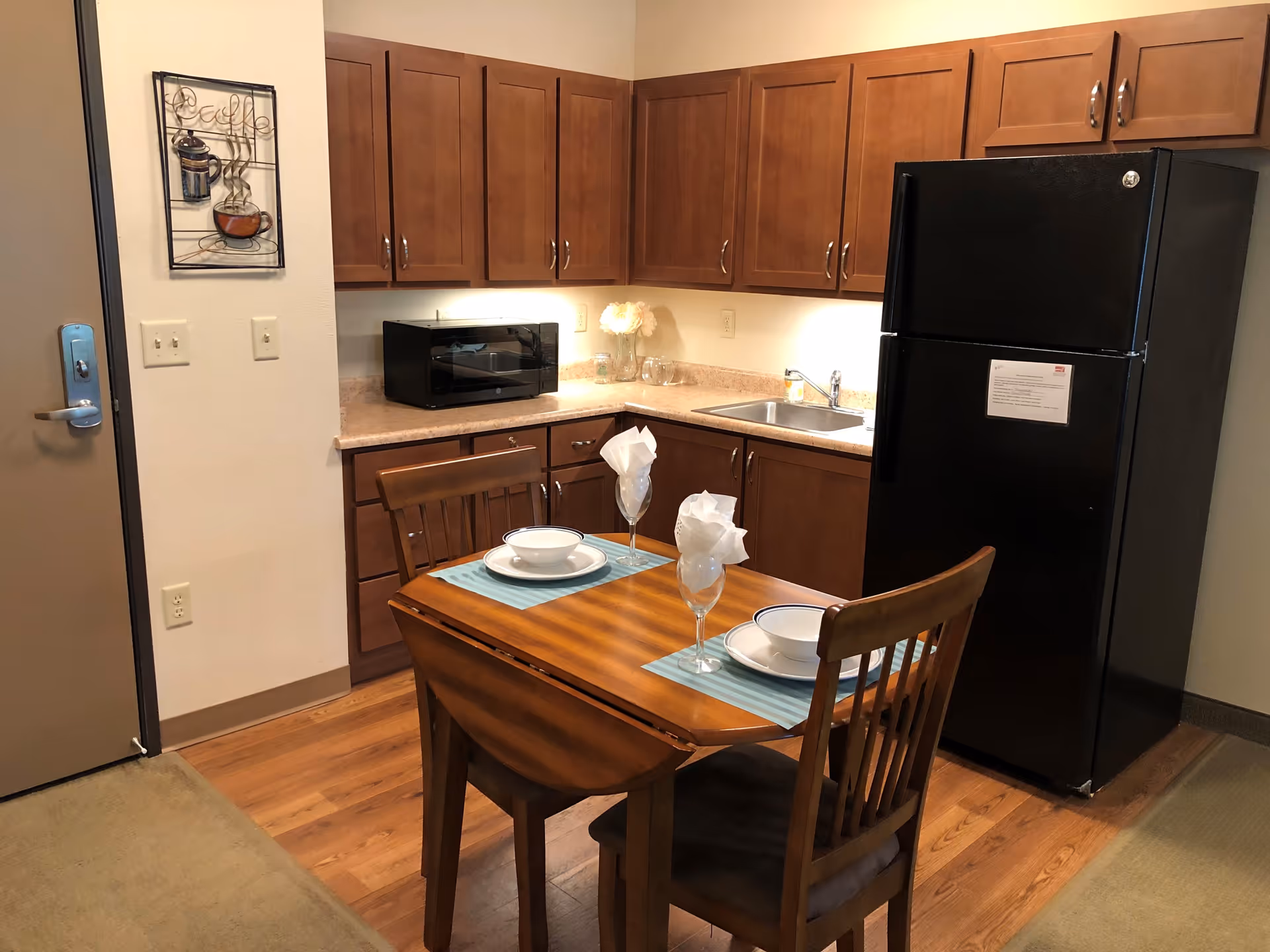 A small kitchen area with wooden cabinets, a black refrigerator, a microwave on the counter, and a sink. In front of the kitchen is a wooden dining table set for two with plates, bowls, and glasses with napkins. The floor is wood, and there is a coffee-themed wall decoration near the door.
