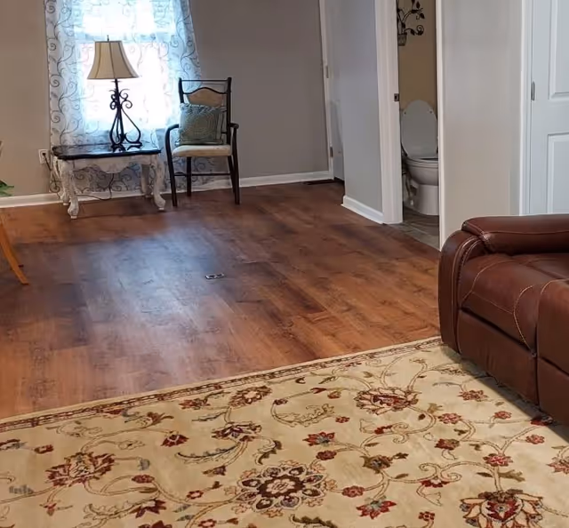 Interior view of a senior living facility room with wooden flooring, a patterned area rug, a brown leather recliner chair, a small table with a lamp, and a chair near a window with sheer curtains. A bathroom with an open door showing a toilet is visible in the background.