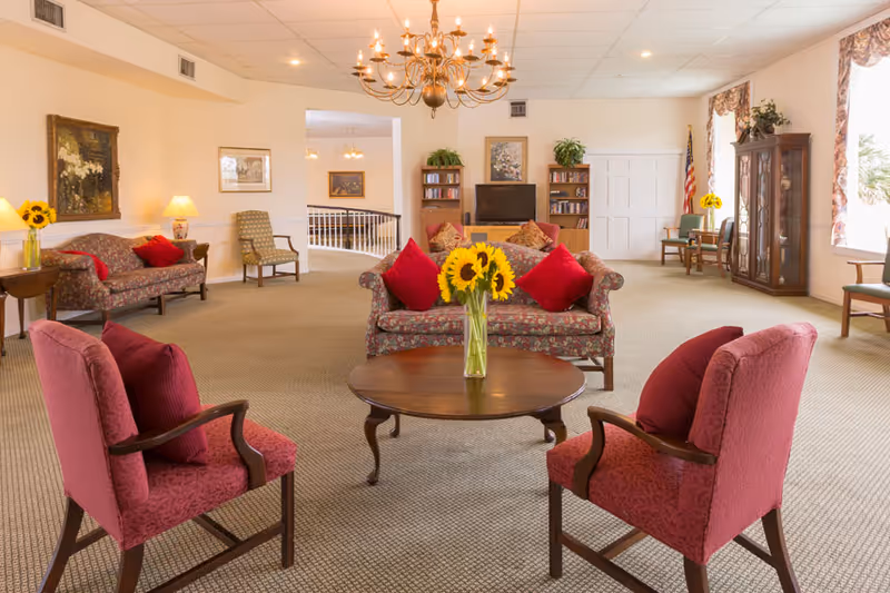 A spacious and well-lit living room area with floral patterned sofas and red cushions, two red upholstered armchairs around a wooden coffee table with a vase of sunflowers, a chandelier hanging from the ceiling, bookshelves with a TV, framed paintings on the walls, and large windows with floral curtains letting in natural light.
