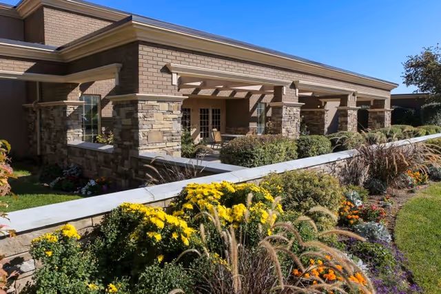Exterior view of a single-story building with stone and brick facade, featuring a covered patio area with columns. In the foreground, there is a well-maintained garden with various colorful flowers and ornamental grasses under a clear blue sky.