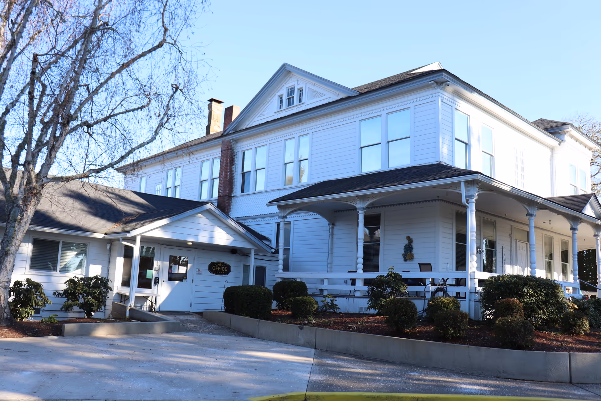 Exterior view of a large white two-story building with a wraparound porch and multiple windows. There is a small entrance area with a sign that reads 'Office' and some bushes and trees around the building. The sky is clear and blue.