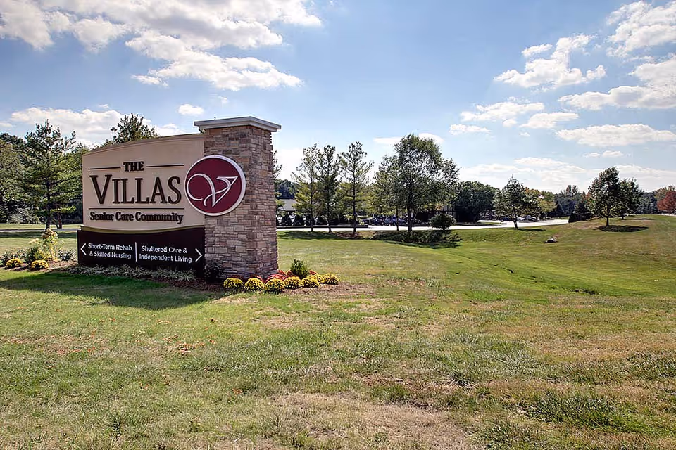 Outdoor view of a large sign for The Villas Senior Care Community on a grassy area with trees and a partly cloudy sky in the background.