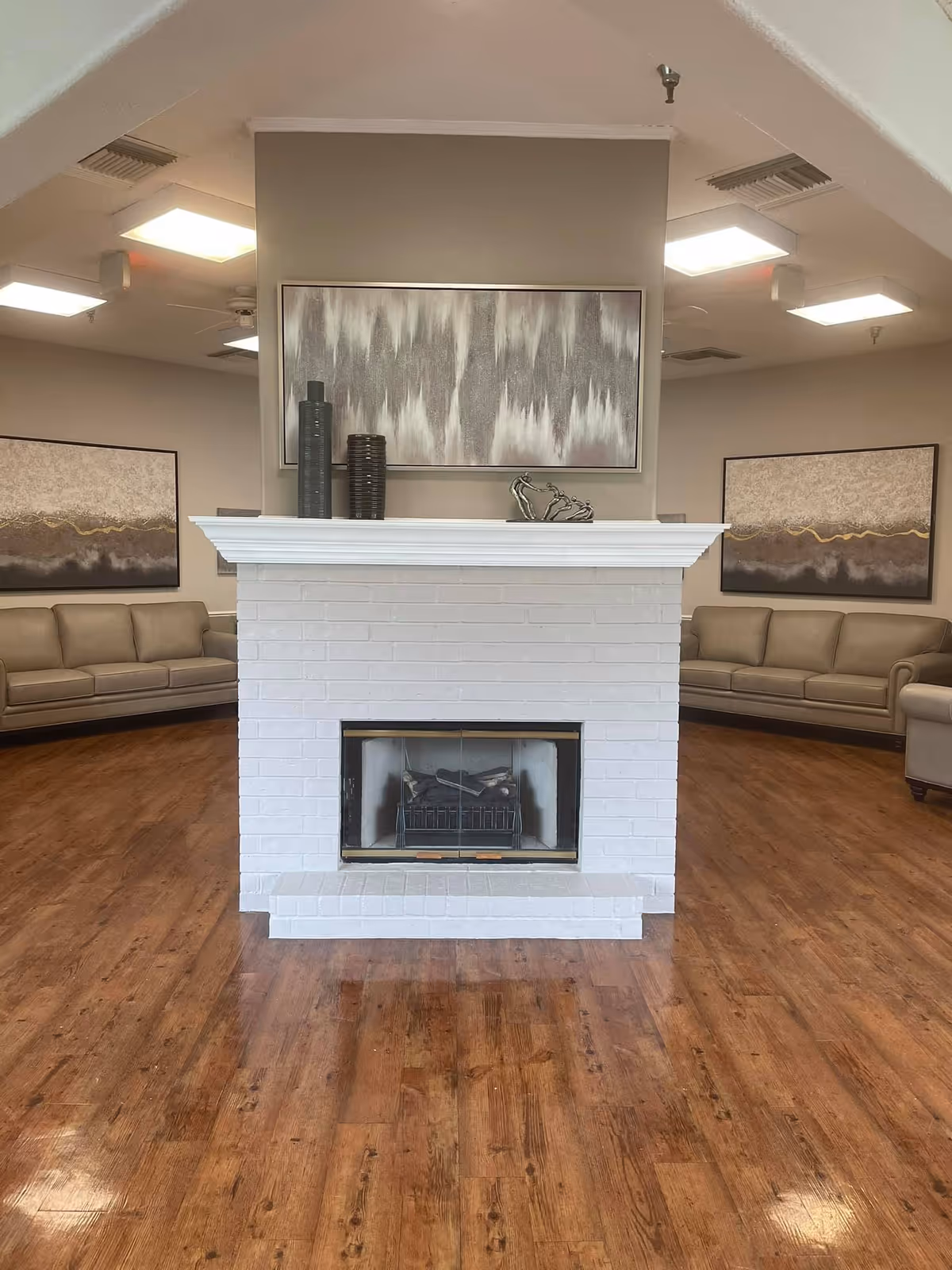 A spacious common area with a white brick fireplace in the center. On the fireplace mantel are decorative vases and a small sculpture. Behind the fireplace, there are two beige sofas arranged against the walls, each with abstract artwork hanging above them. The room has wooden flooring and ceiling lights.