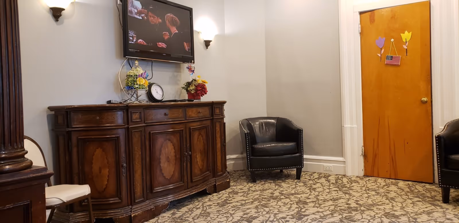 A cozy room with a patterned carpet, two black leather armchairs, a wooden cabinet with decorative items including a clock and flowers, and a wall-mounted TV showing a scene from a show or movie. The room has beige walls, two wall sconces, and a wooden door decorated with paper flowers and a small American flag.
