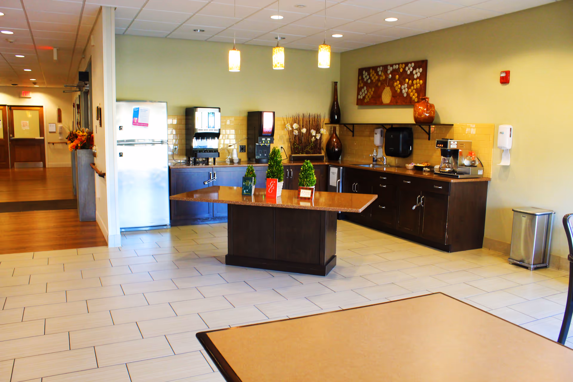 Communal kitchen and coffee station with an island, refrigerator, beverage machines, cabinets and seating in a senior living facility.