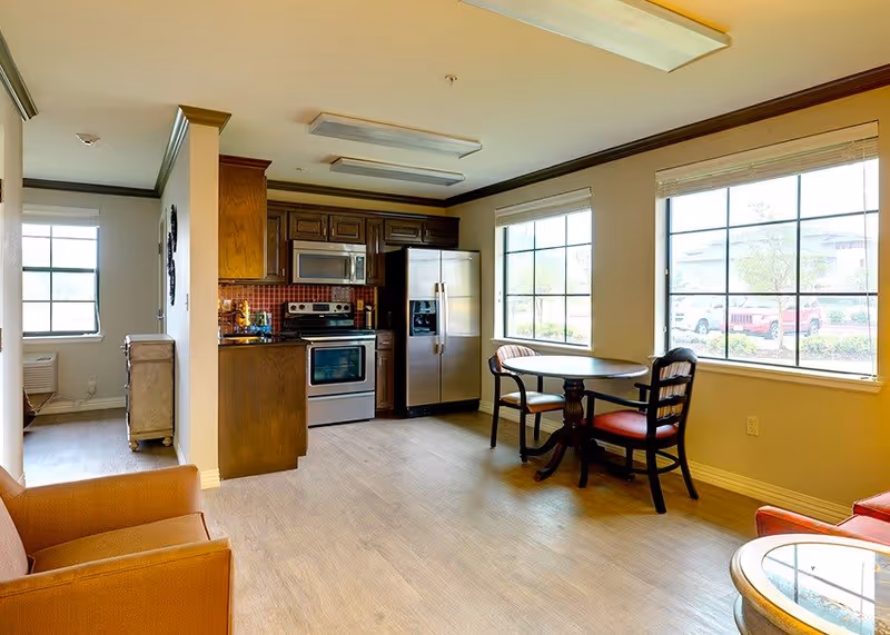 Open-plan senior living interior with a small kitchen featuring stainless-steel appliances, a round dining table by large windows, and armchairs.