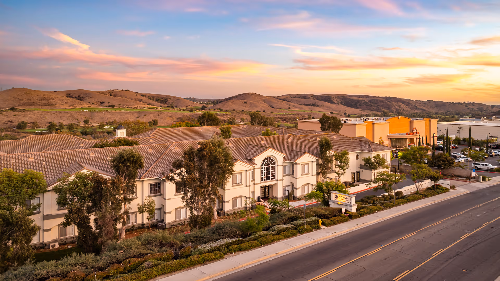 Aerial view of Ivy Park at Mission Viejo senior living facility at sunset, showing a large two-story building with a tiled roof, surrounded by trees and landscaping, with hills in the background and a road in the foreground.
