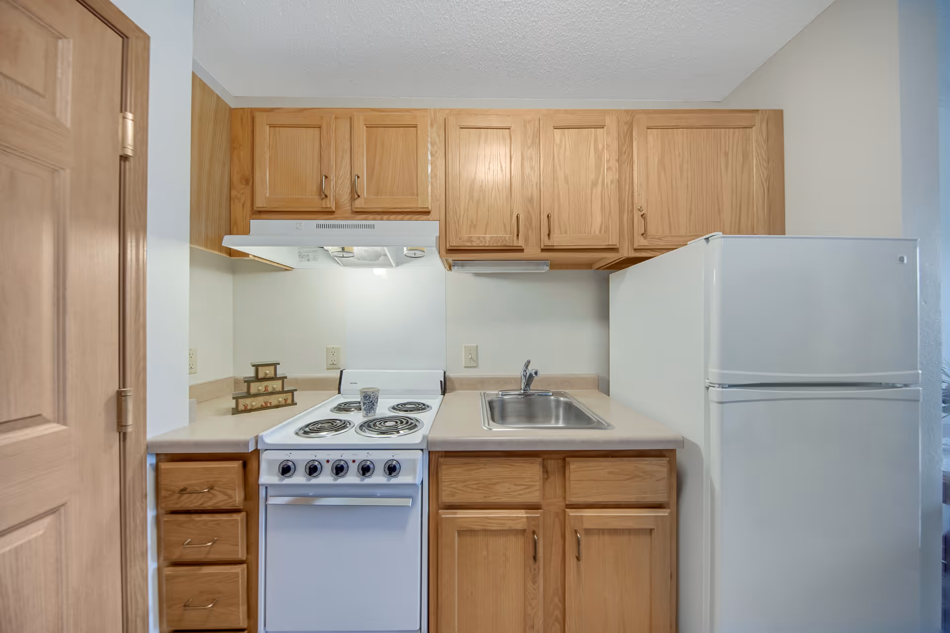Small kitchen area with wooden cabinets, a white electric stove with four coil burners, a stainless steel sink, and a white refrigerator. The countertop is beige, and there is a wooden door on the left side.