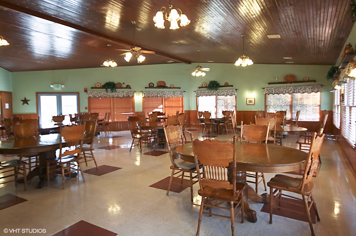 Large dining hall with round wooden tables and chairs beneath a wood-paneled ceiling and ceiling fans.