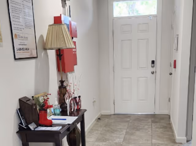 A hallway with a white front door at the end, a small table on the left side holding a lamp, decorative items including a red boot and the word 'JOY', and a framed poster on the wall above the table.