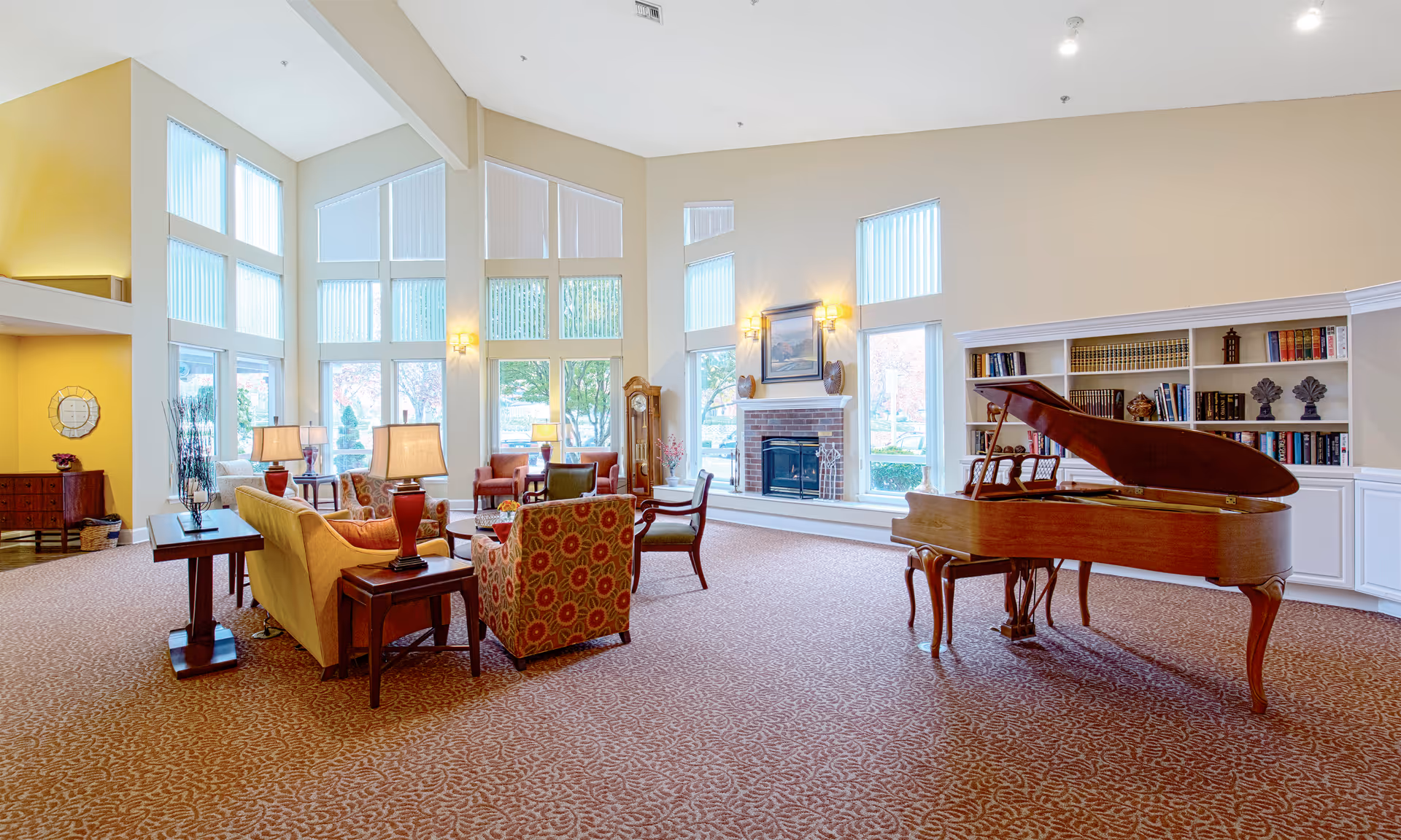Spacious senior living lounge with a grand piano, fireplace, tall windows, and arranged seating areas.
