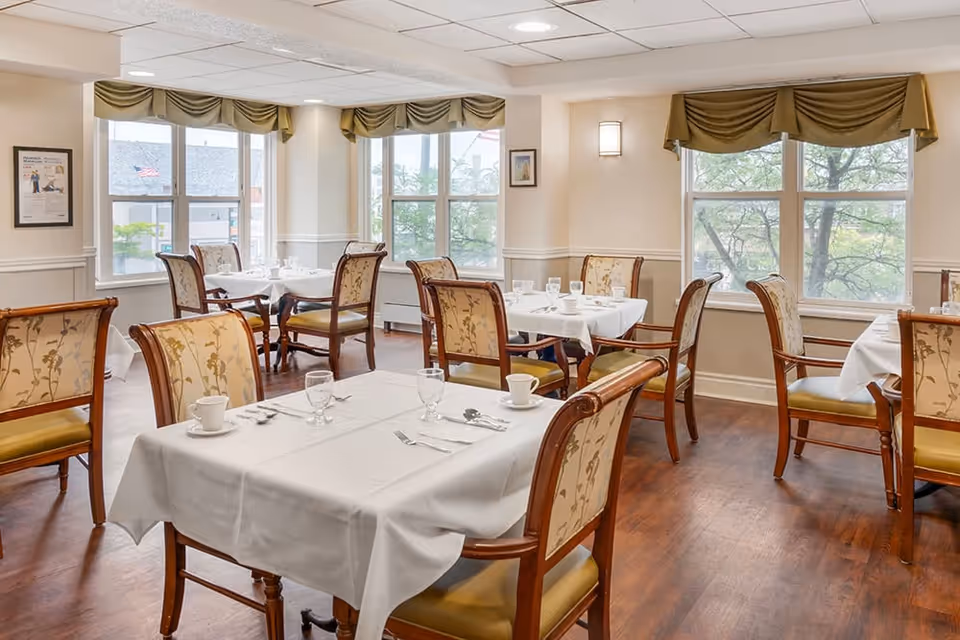 A bright dining room with several tables covered in white tablecloths, each set with cups, glasses, and silverware. The room has large windows with green valances, wooden chairs with floral upholstery, and wooden flooring.