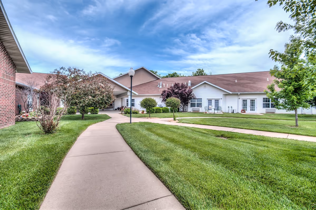 A paved walkway curves through a well-maintained green lawn with small trees and shrubs, leading to a single-story building with white siding and a brown roof under a partly cloudy blue sky.