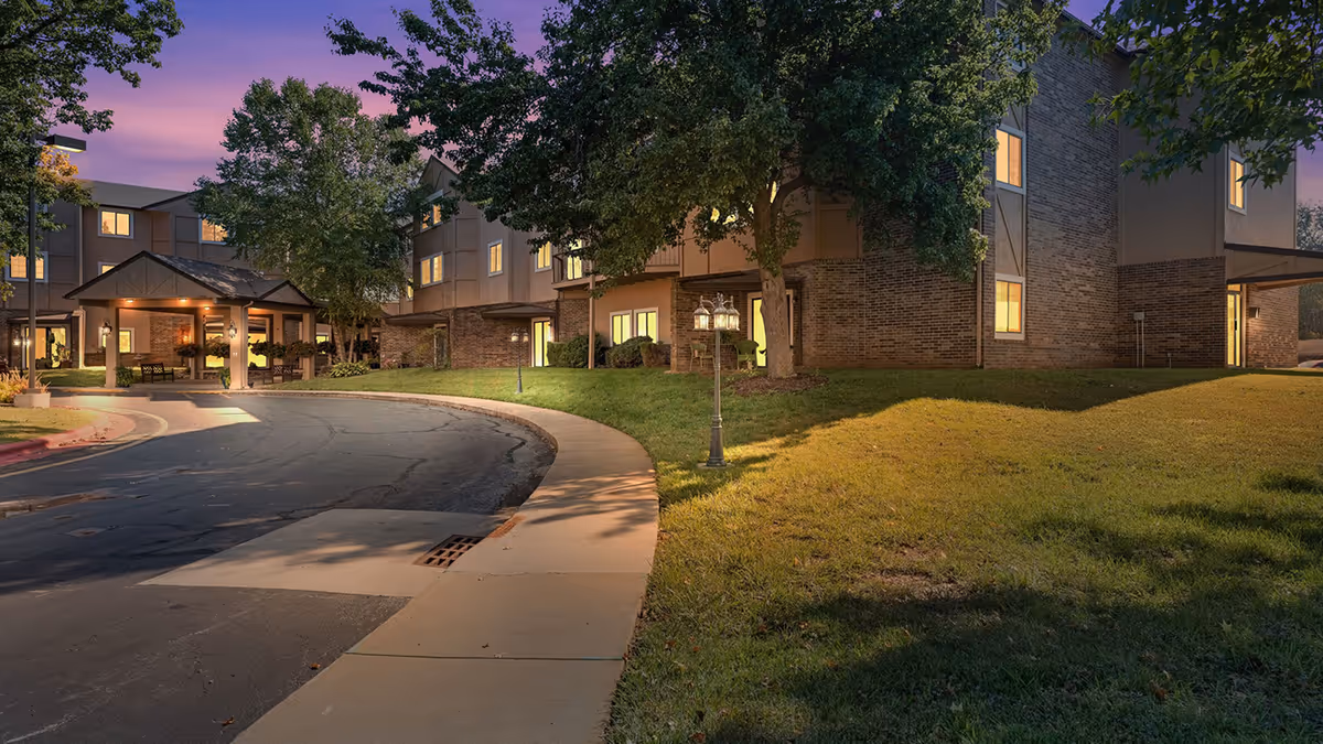 Exterior front entrance of a senior living building at dusk with a covered drop-off, curved driveway, lit windows, and trees.