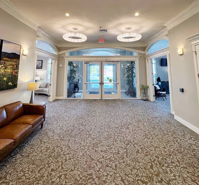 Spacious and well-lit entrance area of an assisted living facility with double glass doors, patterned carpet, a brown leather couch on the left, wall-mounted lights, and modern circular ceiling lights. There are large windows and plants near the doors, and adjacent rooms visible on both sides.