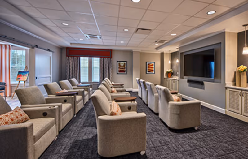 Comfortable senior living media room with two rows of gray recliners facing a large wall-mounted TV.