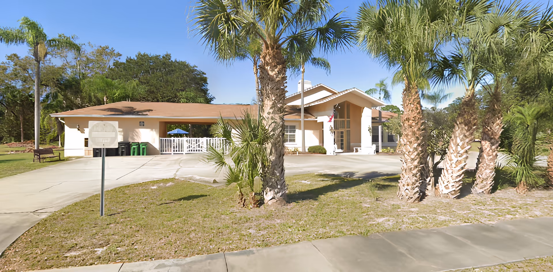 Exterior view of a single-story building with a brown roof and beige walls, surrounded by palm trees and green grass under a clear blue sky. A driveway leads up to the entrance, which has a covered porch area with white pillars.
