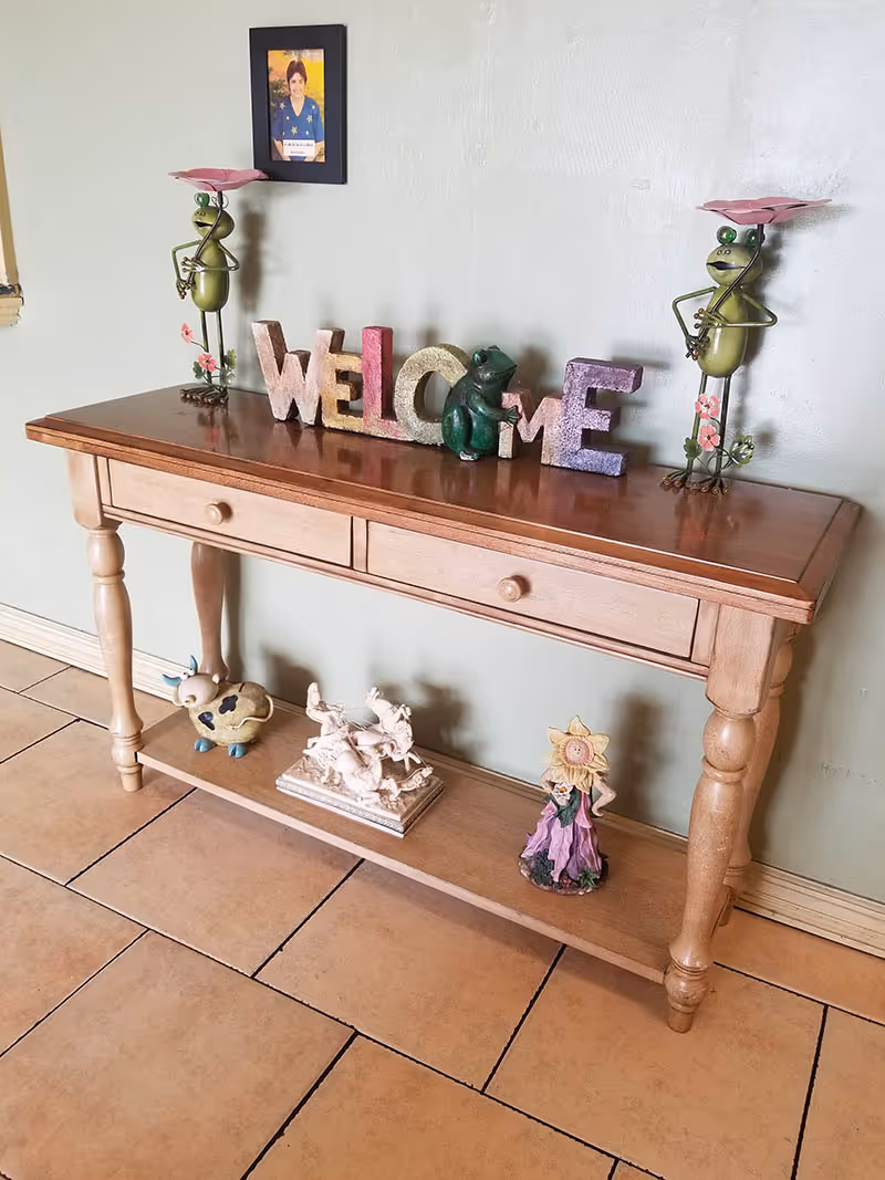 A wooden console table with two drawers and a lower shelf, decorated with colorful 'WELCOME' letters and a small green frog figurine on top. Two metal frog figurines holding pink flowers stand on either side of the letters. On the lower shelf, there is a ceramic cow, a white sculpture of horses, and a figurine of a girl with a sunflower hat. A framed photo hangs on the wall above the table.