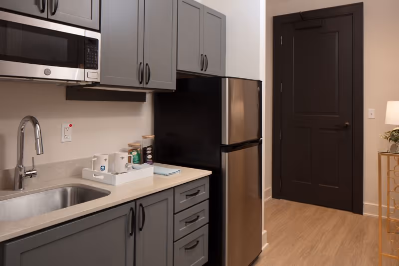 Small modern kitchenette with gray cabinets, stainless steel sink and fridge beside a dark entry door.