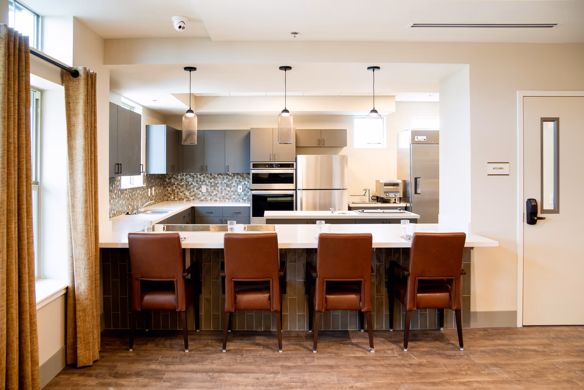 Modern kitchen area with a long white countertop and four brown leather chairs. The kitchen features gray cabinets, stainless steel appliances including a refrigerator and oven, a mosaic tile backsplash, and three pendant lights hanging from the ceiling. There is a window with beige curtains on the left and a door labeled 'Kitchen' on the right.
