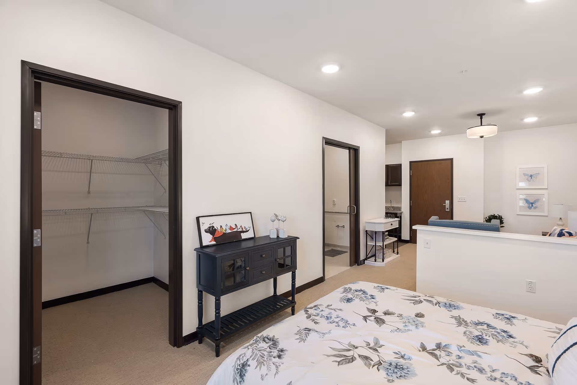 Bright, modern bedroom in a senior living apartment showing a bed with floral bedding, an open closet, a black console table, and a view into the entry and living area.