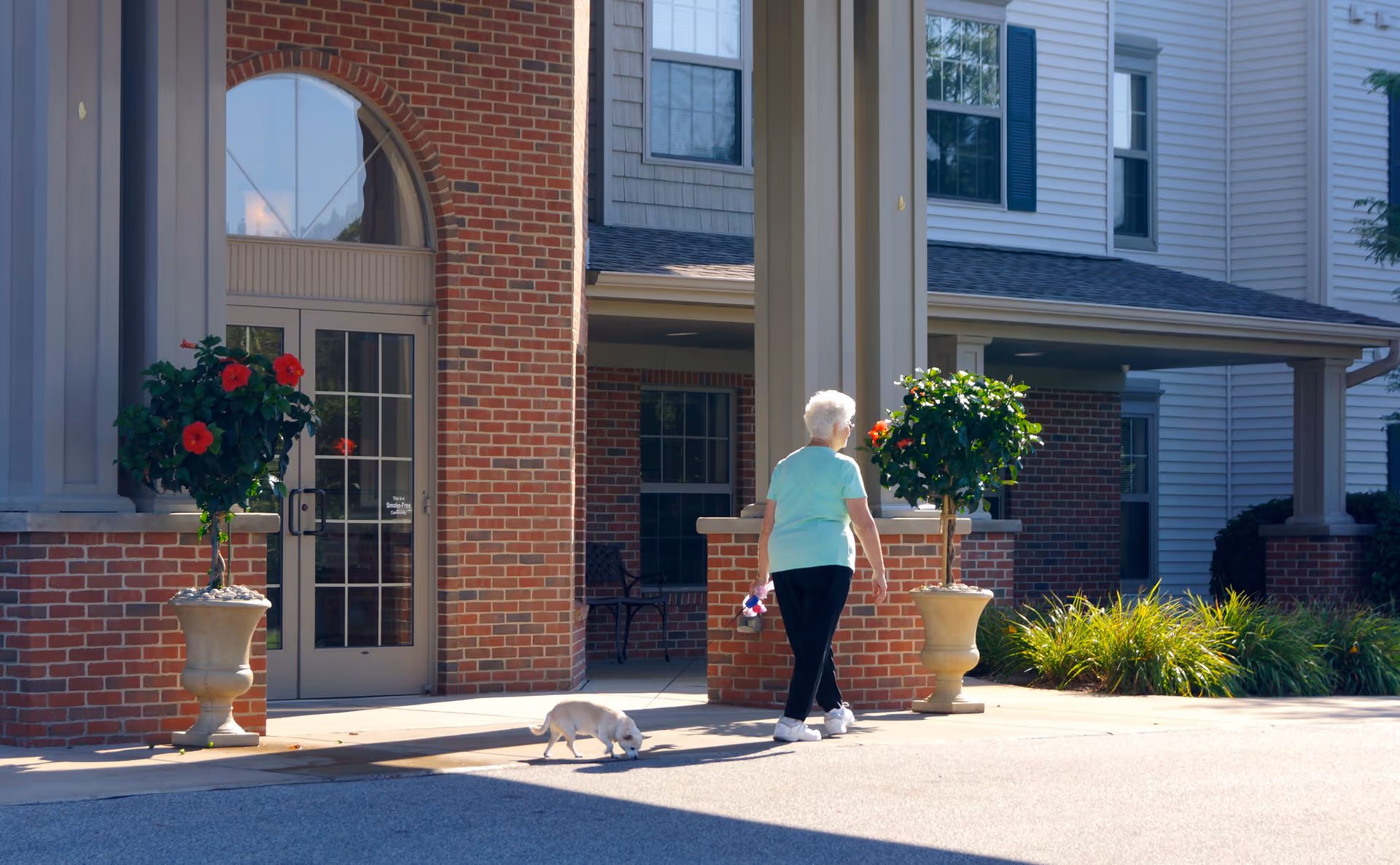 An elderly woman walking a small white dog outside the entrance of a senior living facility with brick and siding exterior, large potted plants, and a glass door.