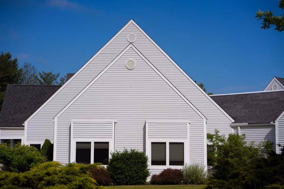 Exterior view of a white building with a steep triangular roof, surrounded by green bushes and trees under a clear blue sky.