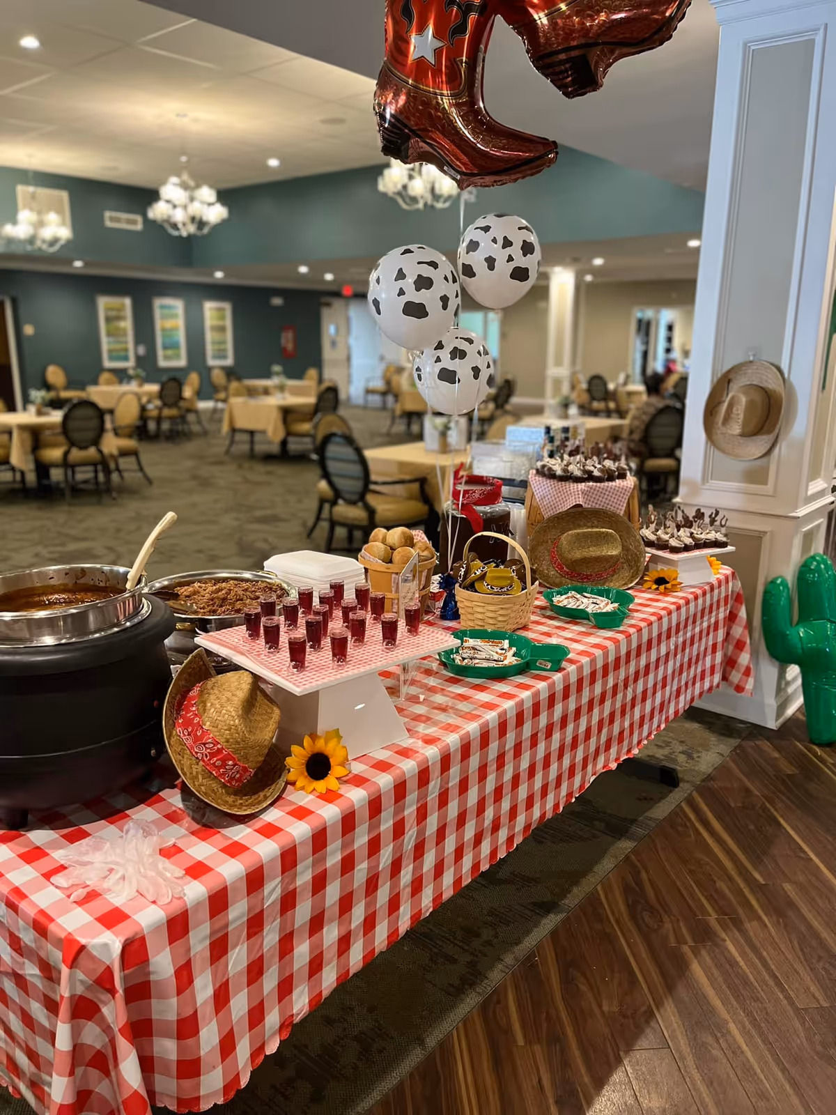 A festive buffet table covered with a red and white checkered tablecloth in a dining area. The table is decorated with cowboy hats, a sunflower, cow-print balloons, and an inflatable green cactus. Various food items including small glasses of a red drink, rolls, and desserts are arranged on the table. In the background, there are multiple round dining tables with chairs and chandeliers hanging from the ceiling.