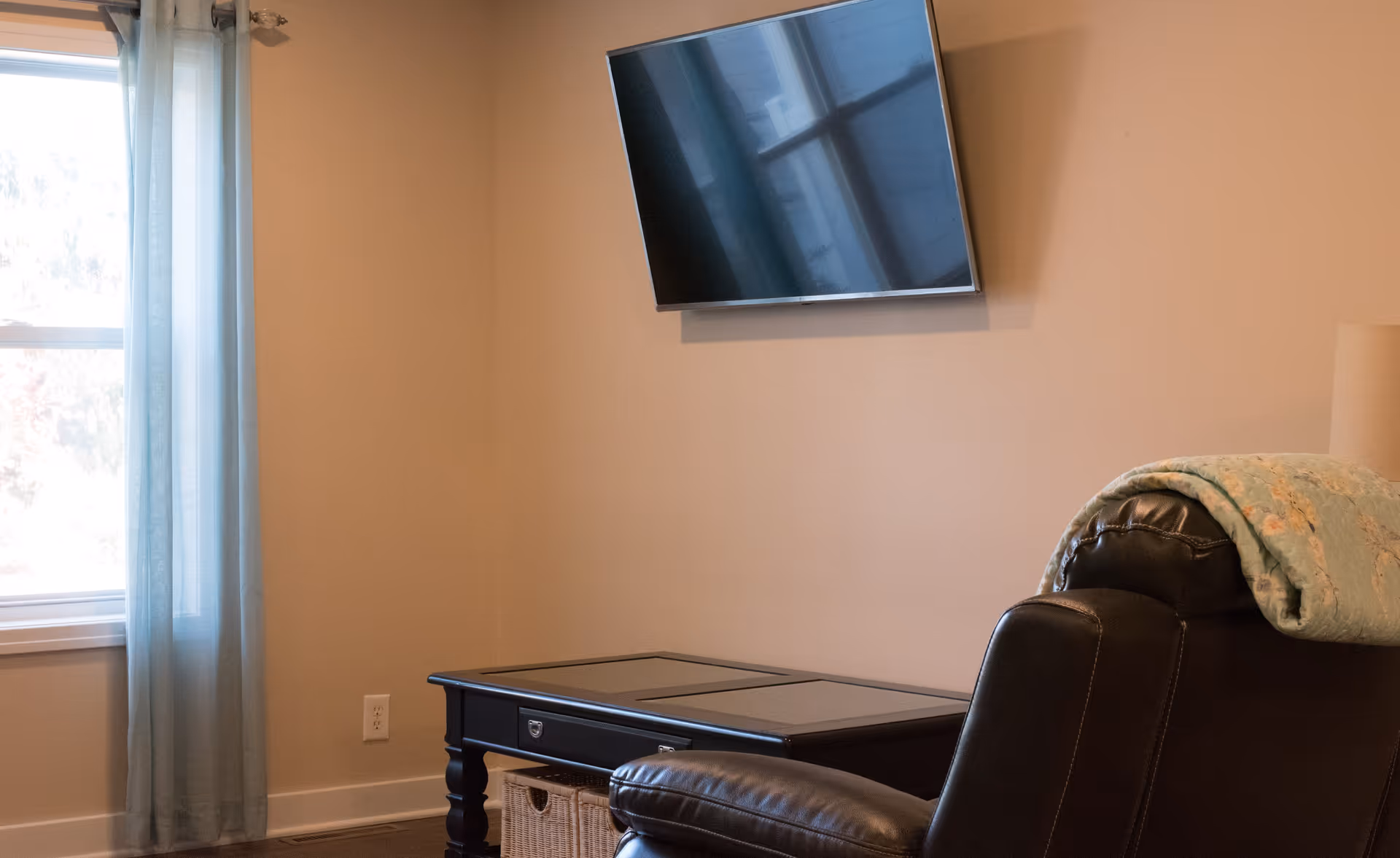 A cozy living room corner with a wall-mounted flat-screen TV, a dark wooden table with a wicker basket underneath, a leather recliner chair with a folded blanket draped over the back, and a window with light blue curtains letting in natural light.