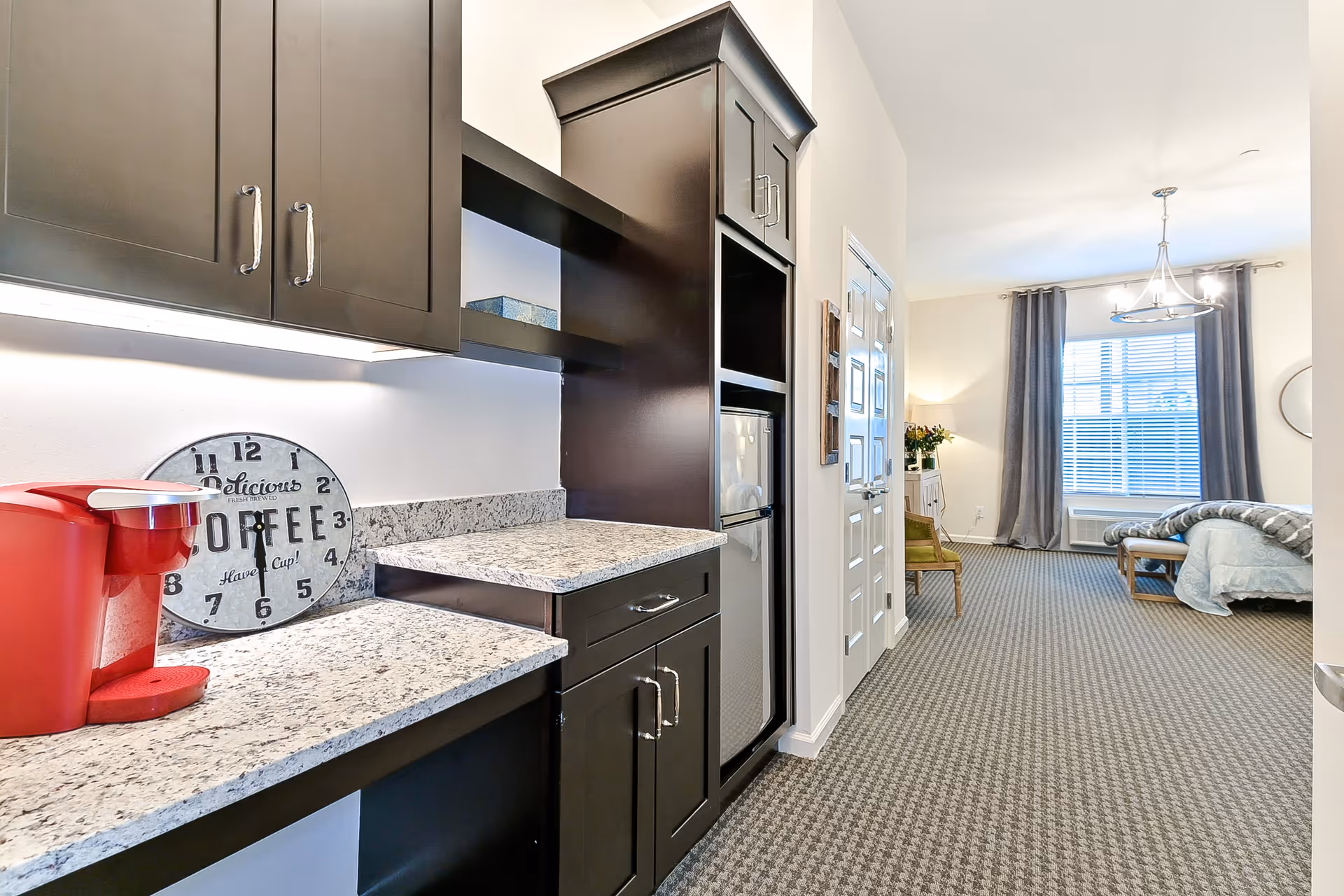 Small kitchenette with dark cabinets and granite countertops opening into a bright studio living/sleeping area with a bed by a large window.