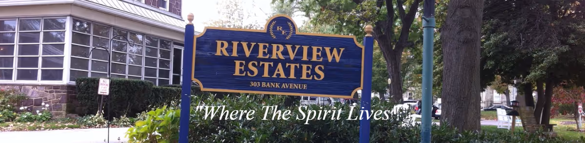 Blue and gold sign for Riverview Estates at 303 Bank Avenue with the tagline 'Where The Spirit Lives' in front of a building with large windows and surrounded by greenery and trees.