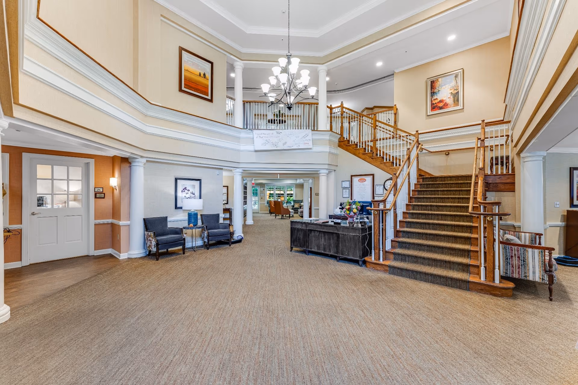 Spacious and well-lit senior living facility lobby with beige carpet, a wooden staircase with carpet runner, white columns, and a chandelier hanging from the ceiling. There are two blue chairs with a small table and lamp between them on the left side, a dark reception desk with flowers on the right, and framed artwork on the walls. The upper level has a railing overlooking the lobby area.