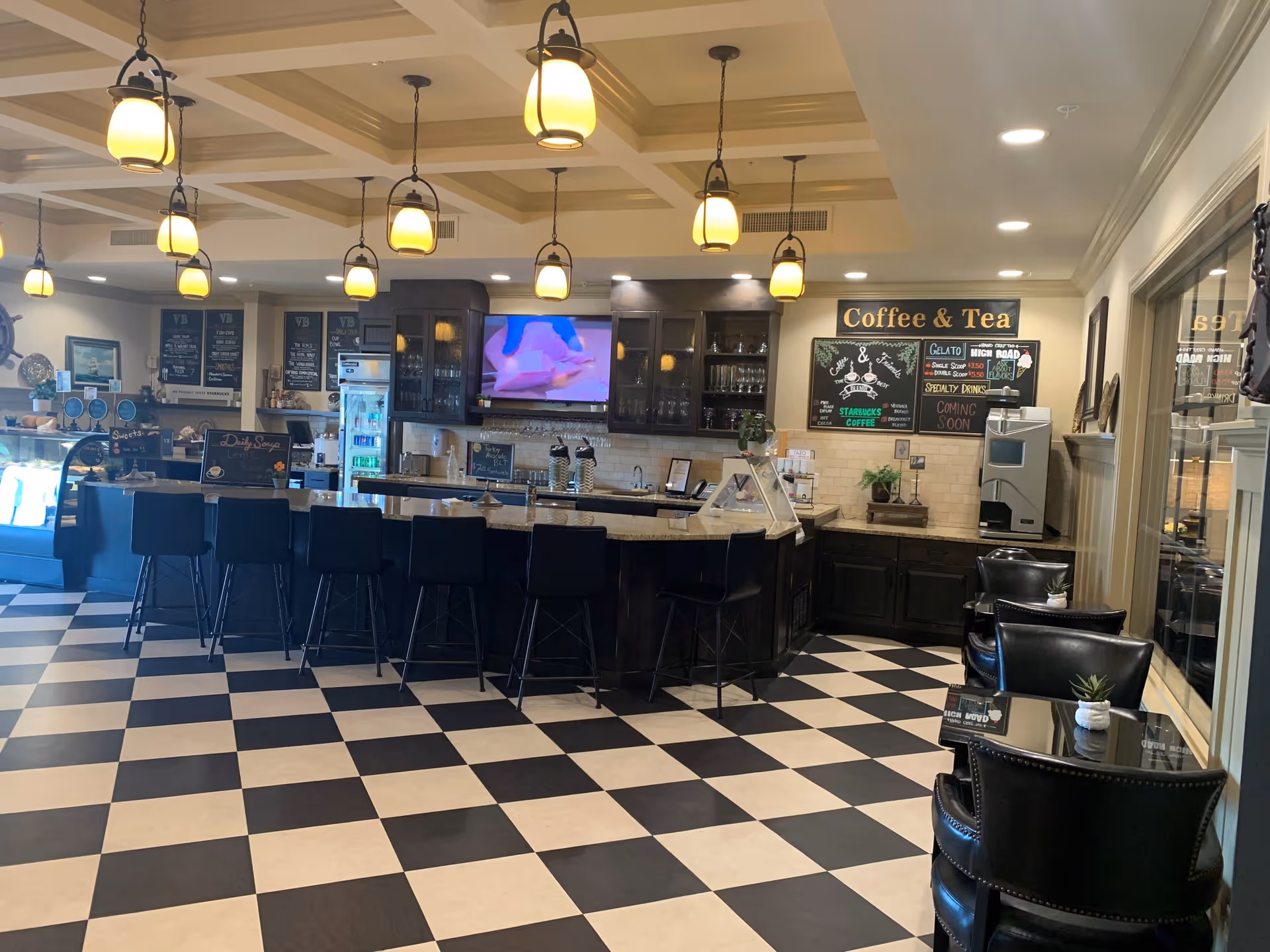 Interior view of a coffee and tea bar area with a black and white checkered floor, a long counter with black bar stools, hanging pendant lights, a menu board on the wall, and small tables with black chairs and potted plants.