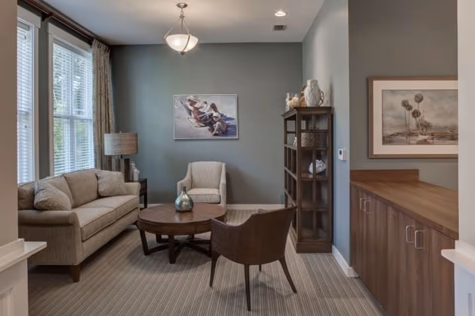 A cozy living room with a sofa, two armchairs, round coffee table, bookshelf, and framed artwork on muted green walls.