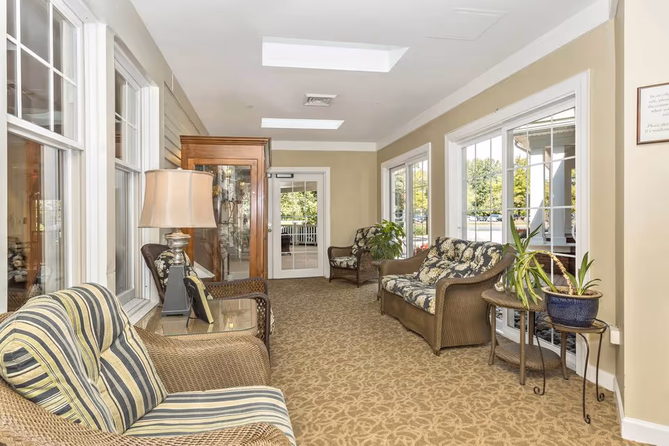 A bright and cozy sitting area with wicker furniture including a striped cushioned chair, a floral cushioned loveseat, and a matching armchair. There is a glass-top side table with a lamp and picture frame, a wooden cabinet, and several potted plants. Large windows line the walls, letting in natural light and providing views of the outdoor greenery.