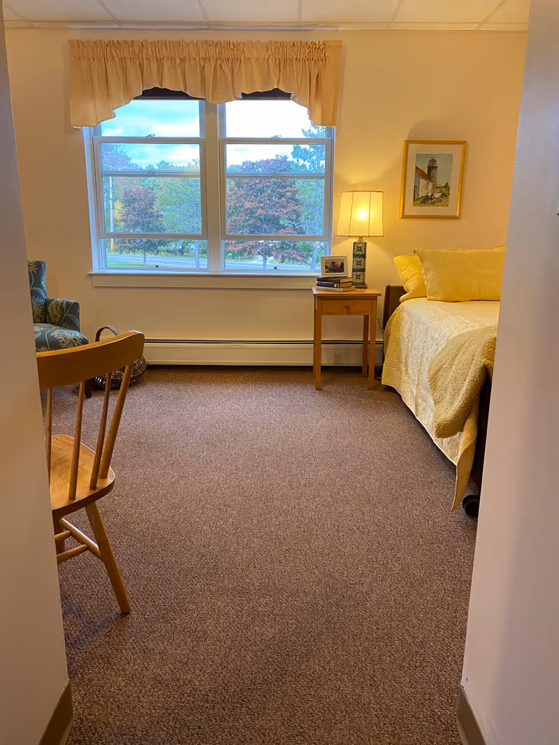 Small bright assisted-living bedroom with a bed, nightstand and lamp beside a large window overlooking trees.