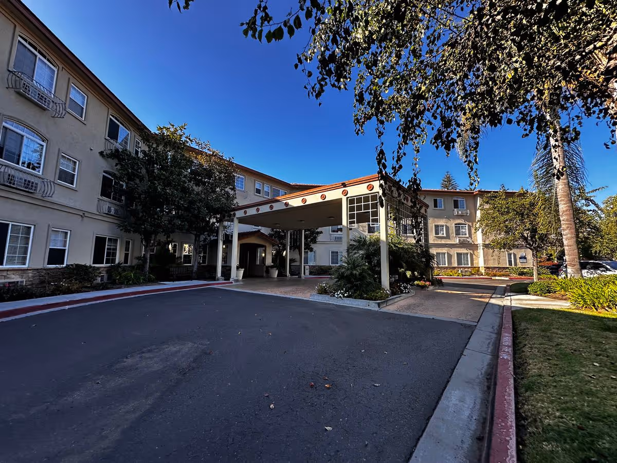 Driveway and covered porte-cochère at the front of a multi-story residential building with landscaping under a clear blue sky.