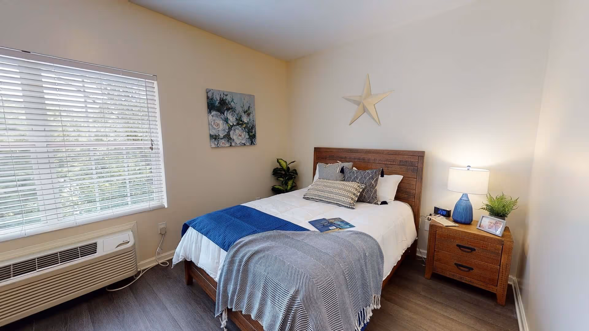 A cozy bedroom with a wooden bed frame and white bedding accented by blue and gray throw blankets and pillows. There is a wooden nightstand with a blue lamp, a small plant, a digital clock, and a framed photo. A large window with white blinds lets in natural light, and a decorative star and floral painting hang on the walls.