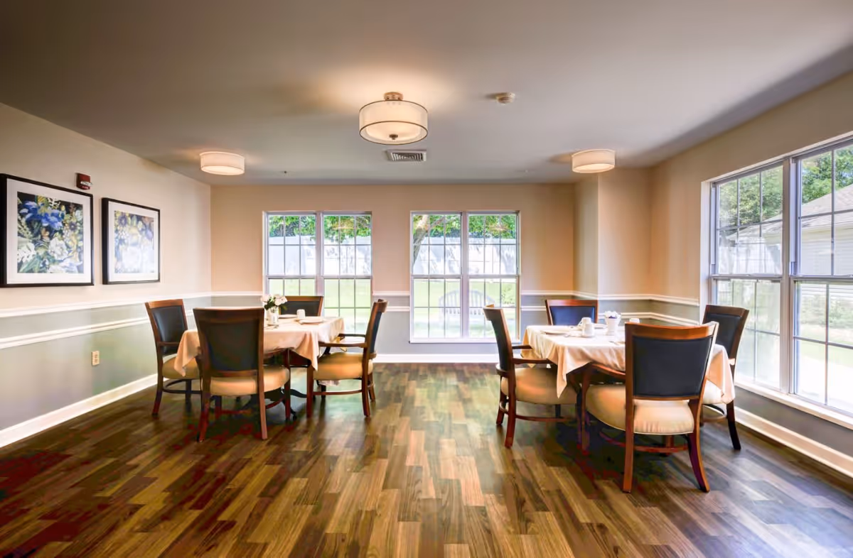 A bright dining room with two round tables covered with white tablecloths, each surrounded by four wooden chairs with cushioned seats. The room has large windows letting in natural light, light-colored walls with white trim, two framed floral paintings on one wall, and wood-patterned flooring.