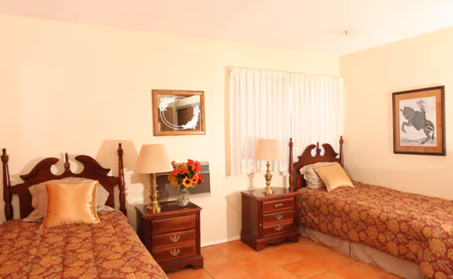 A bedroom with two single beds featuring wooden headboards and matching red and gold patterned bedspreads. Each bed has a gold pillow. Between the beds are two wooden nightstands with brass lamps and a vase of flowers on one. A window with white curtains is centered on the back wall, and a framed picture of a zebra is hung on the right wall.