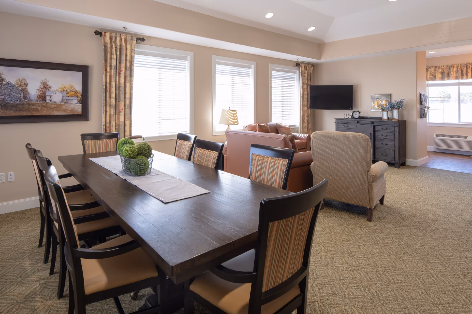 Open common area with a long dining table and chairs in the foreground and a sitting area with sofas, an armchair, and a TV by the windows.
